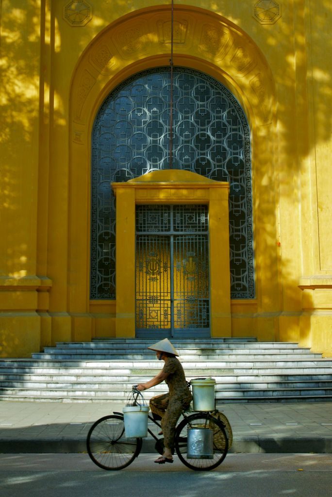 Lokal vietnameser med hatt og bøtter på sykkelstyret sykler forbi en massiv gul bygning med trapper foran